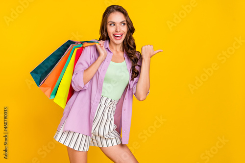 Photo of amazed cheerful young woman hold shopping bags smile sale point finger empty space isolated on yellow color background