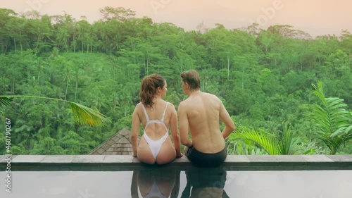 A couple in love, a man in black shorts and a girl in a white swimsuit, are sitting on the edge of the pool with a view of the rainforest and sunset. Male pushes the girl and she falls into the pool.