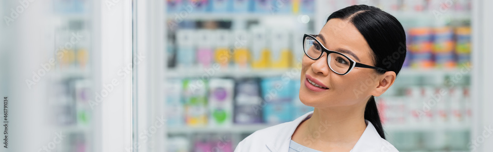 smiling asian pharmacist in glasses looking away in drugstore, banner - obrazy, fototapety, plakaty 