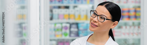 smiling asian pharmacist in glasses looking away in drugstore, banner
