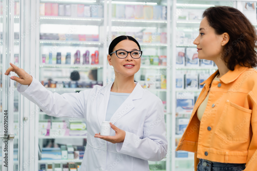 cheerful asian pharmacist in glasses holding bottle with drugs and pointing away near brunette customer in drugstore