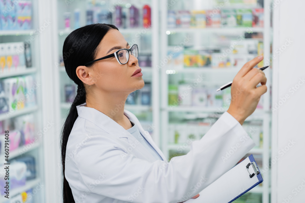 asian pharmacist in glasses holding pen while pointing at drugs