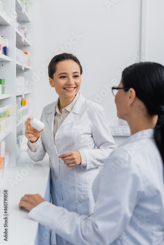 cheerful pharmacist in white coat holding bottle with medication near blurred asian colleague in glasses