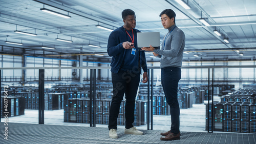 Two Multiethnic Male Data Center System Administrators and IT Specialists Talk, Use Laptop. Information Technology Engineers work on Cyber Security Network Protection in Cloud Computing Server Farm.