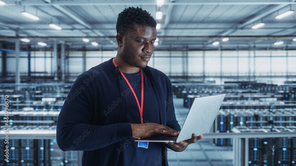 Portrait of a Data Center Engineer Using Laptop Computer. Server Room Specialist Facility with African American Male System Administrator Working with Data Protection Network for Cyber Security. Portrait of a Data Center Engineer Using Laptop Computer. Server Room Specialist Facility with African American Male System Administrator Working with Data Protection Network for Cyber Security.