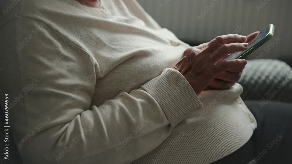 Old woman with wrinkled hands dressed in white blouse scrolls mobile phone sitting in soft armchair watching screen closeup Old woman with wrinkled hands dressed in white blouse scrolls mobile phone sitting in soft armchair watching screen closeup