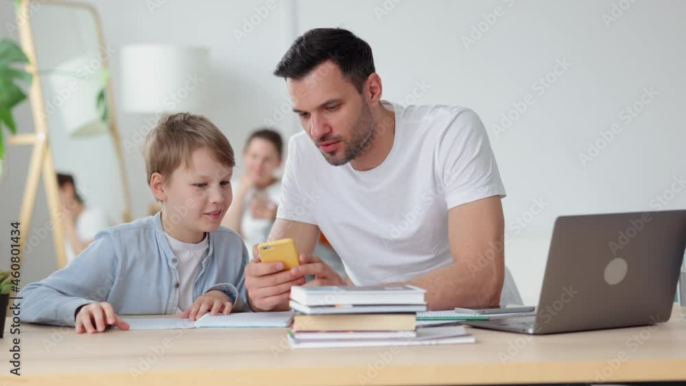 Dad, son using smartphone and sitting at table with laptop in home room spbi. Closeup view of young man, child hold phone in hands and look with smile, talk and sit at desk with computer in interior Dad, son using smartphone and sitting at table with laptop in home room spbi. Closeup view of young man, child hold phone in hands and look with smile, talk and sit at desk with computer in interior