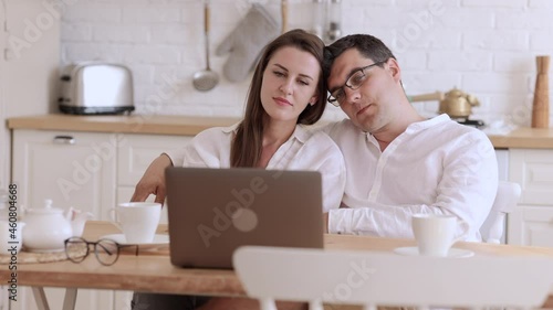 Young couple hugging and usig laptop while sitting at table in home room spbi. Closeup view of beautiful woman, man hug and look at computer screen, talk with smiles and sit at desk in light interior photo