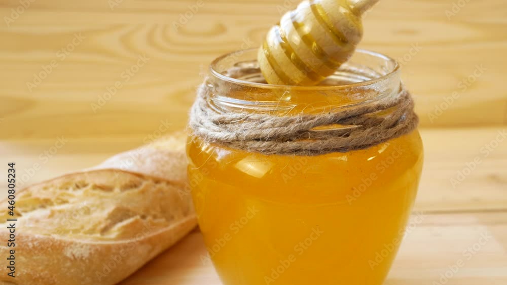 A wooden spoon is stirring thick viscous honey in a glass jar. There is a baguette next to it. Organic honey. natural products and proper nutrition. A wooden spoon is stirring thick viscous honey in a glass jar. There is a baguette next to it. Organic honey. natural products and proper nutrition.