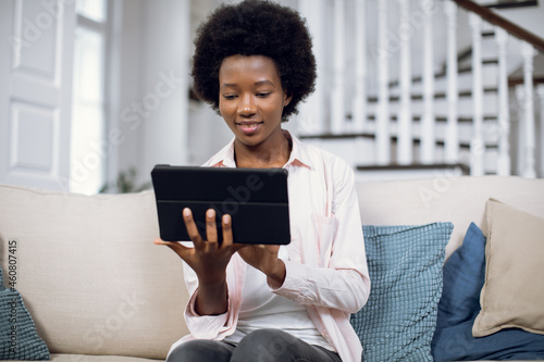 Likable pretty african american woman using digital tablet while resting on comfy couch indoors at home. Young female in casual outfit smiling and looking at digital screen, reading news