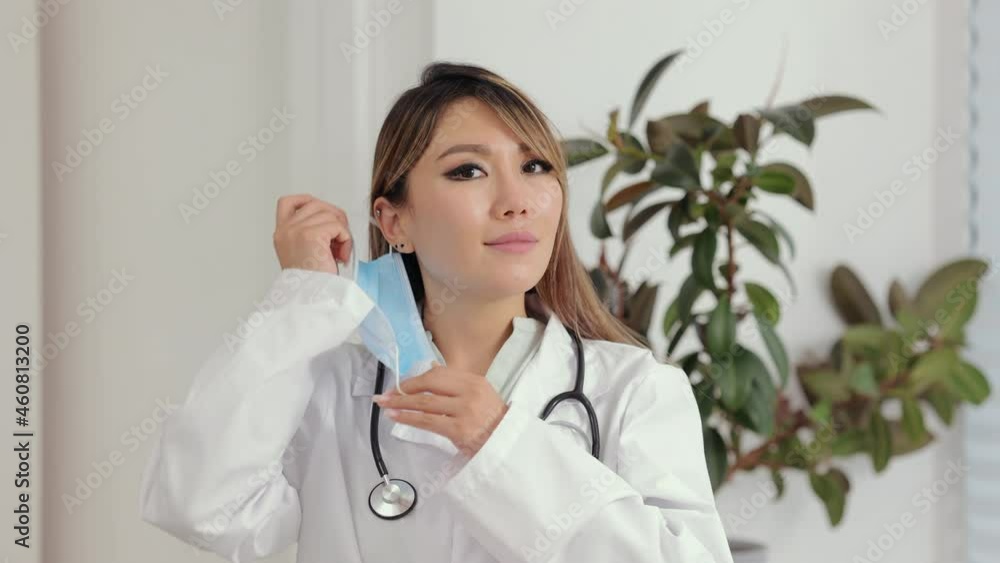 Woman doctor wearing mask on face posing for camera and standing in clinic office spbi. Young beautiful woman takes off her mask and looks with smile, stands in light room. Medical expert poses in Woman doctor wearing mask on face posing for camera and standing in clinic office spbi. Young beautiful woman takes off her mask and looks with smile, stands in light room. Medical expert poses in