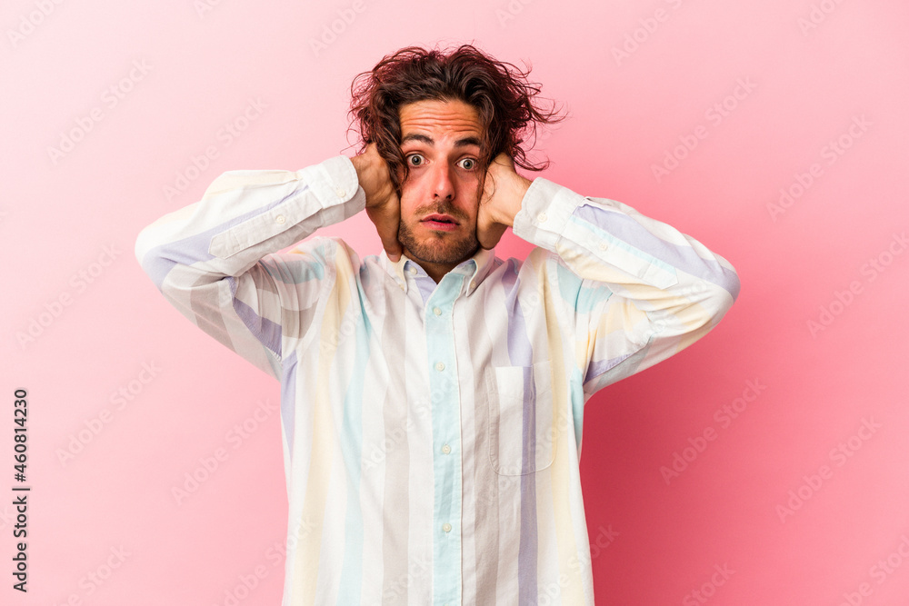 Young caucasian man isolated on pink bakcground covering ears with hands trying not to hear too loud sound.