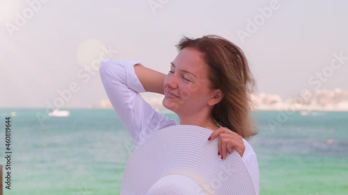 A young woman in a white shirt stands in front of the sea and straightens hair. A girl on vacation enjoying lifestle and freedom on the coast beach. Weekend and holiday on the seashore of blue sea. photo