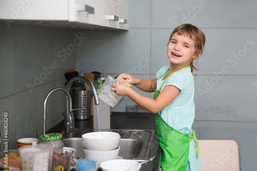 A little cute child girl washing dishes in the white kitchen interior. casual lifestyle photo series in real life interior. Child with helping his parents with housework.
