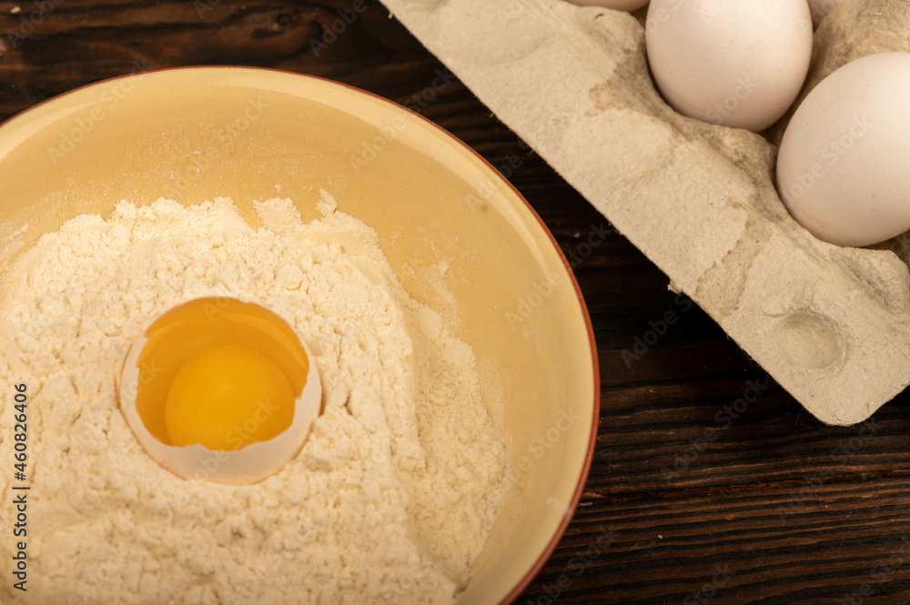 A broken white chicken egg in a bowl of wheat flour and several whole eggs in a tray made of white cardboard, close-up selective focus. A broken white chicken egg in a bowl of wheat flour and several whole eggs in a tray made of white cardboard, close-up selective focus.
