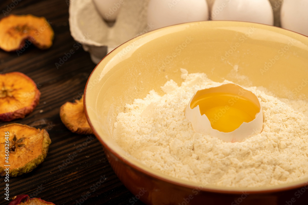 A broken white chicken egg in a bowl with wheat flour, dried apple chips and several whole eggs in a tray made of white cardboard, close-up selective focus. A broken white chicken egg in a bowl with wheat flour, dried apple chips and several whole eggs in a tray made of white cardboard, close-up selective focus.