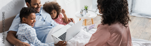 happy african american kids looking at laptop near dad and blurred mom in bedroom  banner