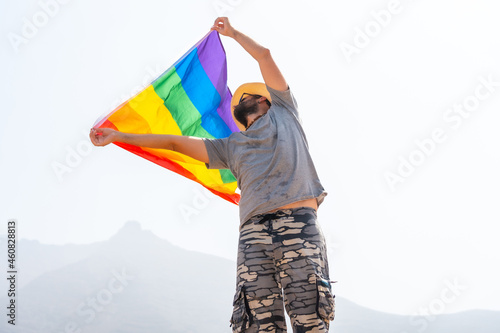 A gay person with a gray t-shirt and white hat waving the LGBT flag in a cloudy sky, symbol of homosexuality