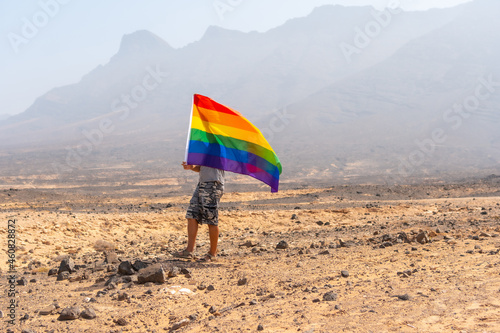 An unrecognizable gay person in a gray t-shirt and black cap with the LGBT flag waving in a desert with the wind, a symbol of homosexuality