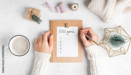 Desktop of woman hands with blank notepad writing 2022 goals text . Flat lay of white working table background with cup of coffee and Christmas decoration. Top view mock up