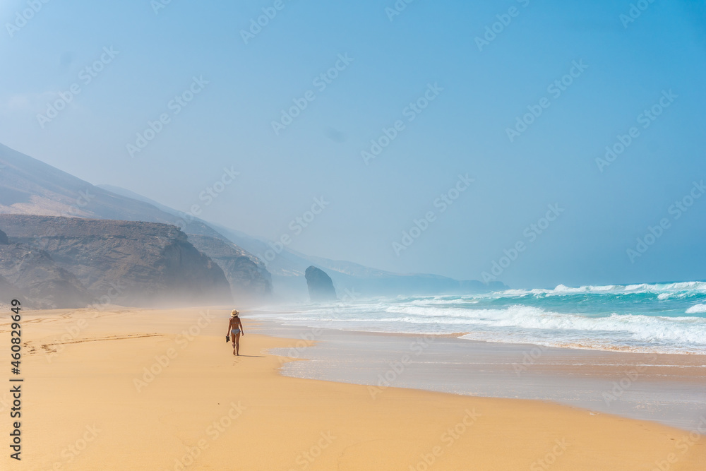 Tourist girl with a hat walking alone on the Cofete beach of the natural park of Jandia, Barlovento, south of Fuerteventura, Canary Islands. Spain