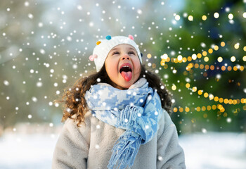 childhood, leisure and winter holidays concept - happy little girl having fun and catching snow by her tongue at winter park