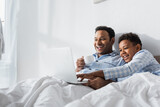 excited african american boy and dad watching comedy on laptop in morning in bed