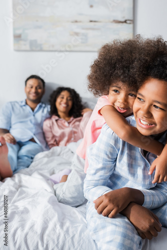 cheerful african american children in pajamas embracing near blurred parents in bedroom