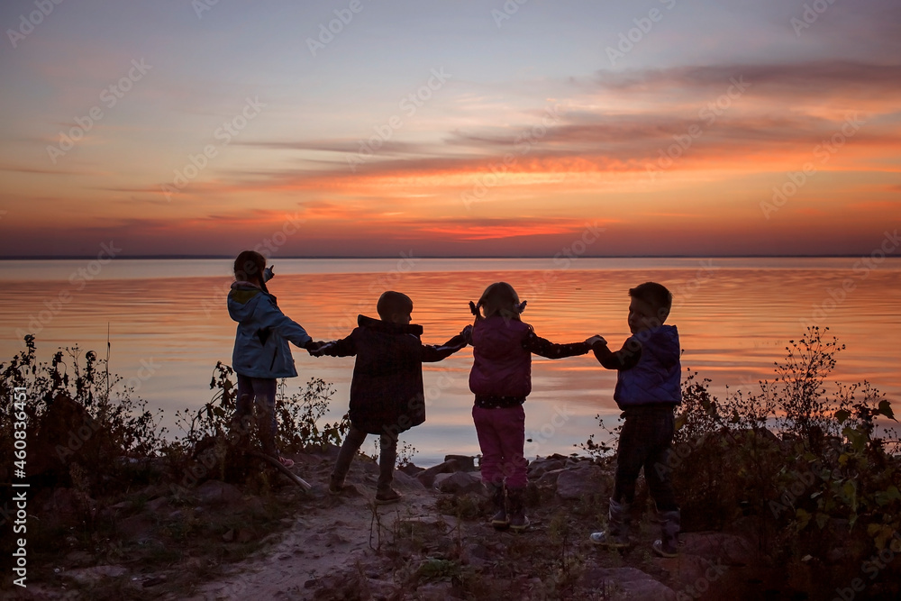 Four kids of different ages stand together holding by hands and look at sun that sets over the sea, trust and friendship, active family weekend. Autumn outside lifestyle, amazing silhouettes