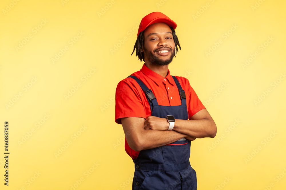 Satisfied handyman wearing overalls standing with crossed arms, looking at camera, profession of service industry, courier delivery. Indoor studio shot isolated on yellow background. Satisfied handyman wearing overalls standing with crossed arms, looking at camera, profession of service industry, courier delivery. Indoor studio shot isolated on yellow background.
