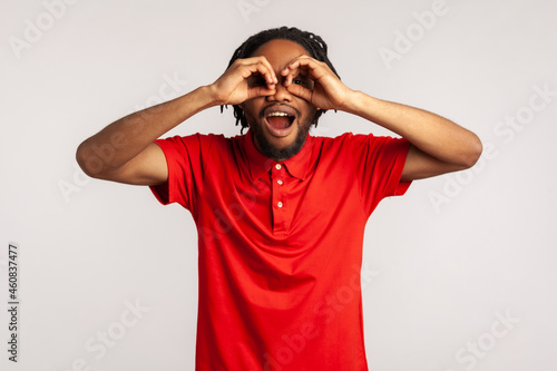 фотографія Amazed young adult man with dreadlocks wearing red casual style T-shirt, looking through fingers shaped like binoculars and expressing excitement