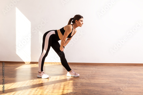 Full length portrait of warming up before training, touching leg with both arms, stretching, wearing black sports top and tights. Indoor studio shot illuminated by sunlight from window.