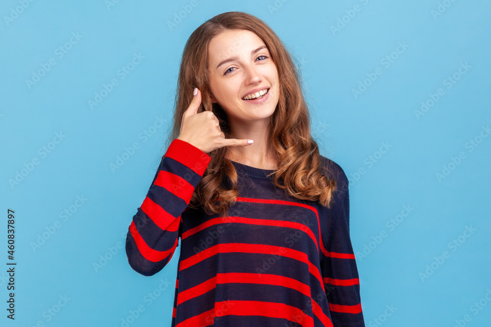 Hey handsome, call me. Smiling woman wearing striped casual style sweater flirting holding fingers near ear showing call gesture, answering call. Indoor studio shot isolated on blue background. Hey handsome, call me. Smiling woman wearing striped casual style sweater flirting holding fingers near ear showing call gesture, answering call. Indoor studio shot isolated on blue background.