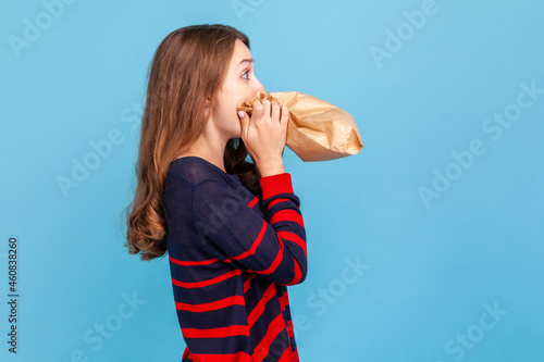 Photography Side view of nervous stressed woman wearing striped casual style sweater, hardly breathing using paper bag, trying to calm down, to improve well-being