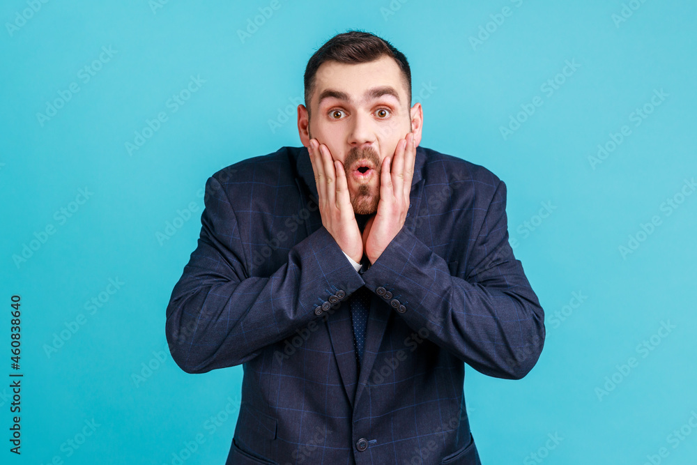 Photo of amazed bearded young adult man in official style suit looks at camera with open mouth, raising palms, being impressed of shocking news. Indoor studio shot isolated on blue background. Photo of amazed bearded young adult man in official style suit looks at camera with open mouth, raising palms, being impressed of shocking news. Indoor studio shot isolated on blue background.
