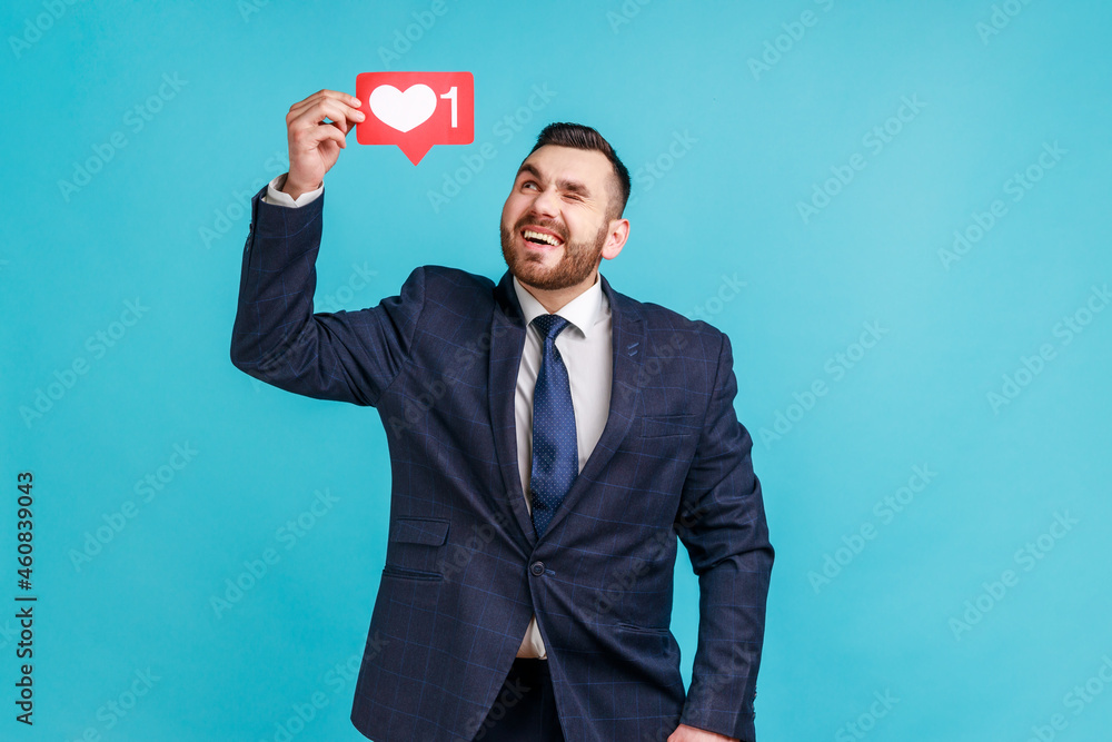 Optimistic bearded man wearing official style suit, holding social network Heart Like icon over head, looking at symbol and winking. Indoor studio shot isolated on blue background. Optimistic bearded man wearing official style suit, holding social network Heart Like icon over head, looking at symbol and winking. Indoor studio shot isolated on blue background.