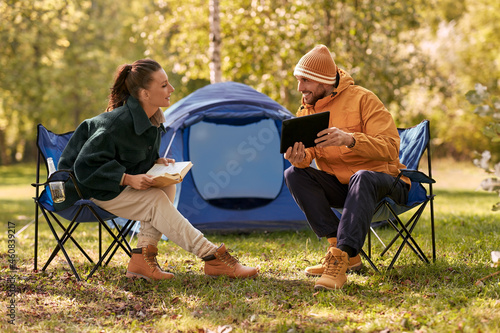 camping, tourism and travel concept - happy couple with tablet pc computer and book resting at tent camp