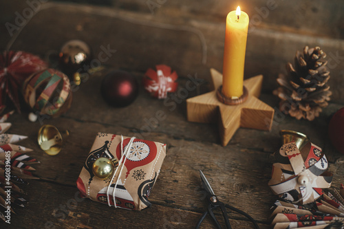 Stylish wrapped Christmas gift box, scissors, candle, red paper stars, golden ornaments on rustic wooden table. Atmospheric image. Merry Christmas and Happy Holidays!