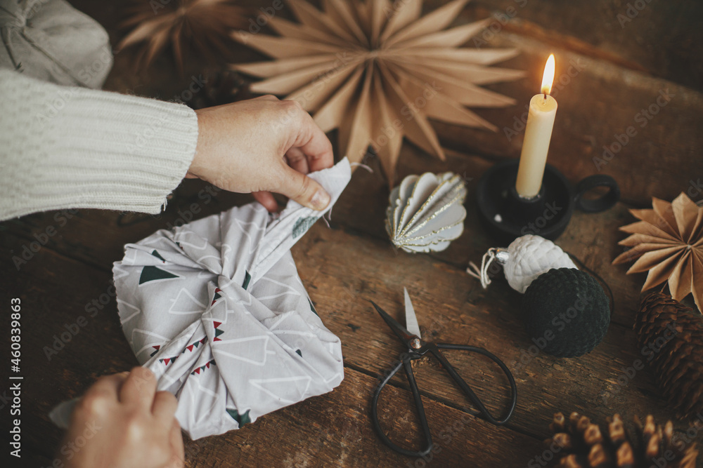 Furoshiki Christmas gift. Hands wrapping christmas gift in modern fabric on rustic wooden table with scissors, craft paper star, candle. Atmospheric moody time, nordic style. Zero waste holiday - obrazy, fototapety, plakaty 