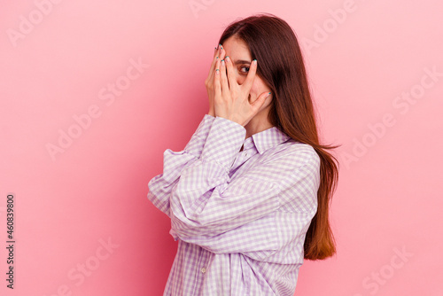 Young caucasian woman isolated on pink background blink through fingers frightened and nervous.