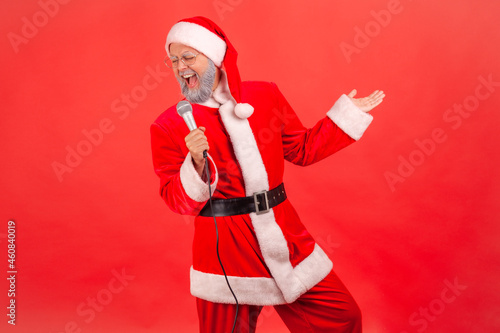 Happy positive elderly man with gray beard wearing santa claus costume standing with microphone in hands, singing songs, celebrating Christmas. Indoor studio shot isolated on red background.