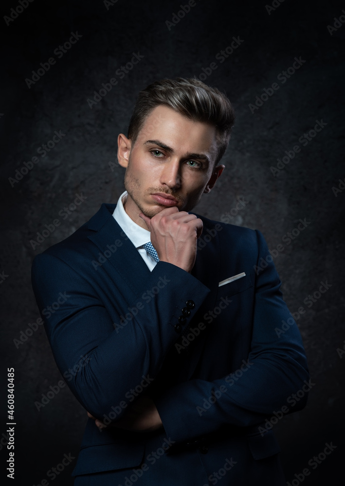 Portrait of a conceived, young, stylish business man, in a blue suit against a dark textured background.