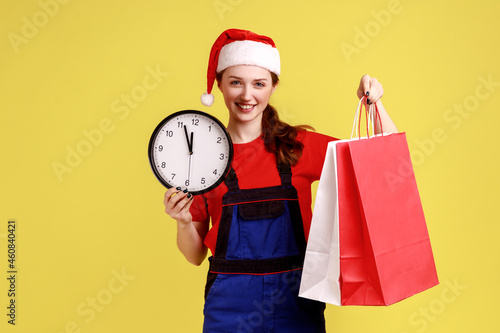 Smiling happy delivery woman holding wall clock and shopping bags, fast delivery for winter holiday, wearing blue overalls and santa claus hat. Indoor studio shot isolated on yellow background.