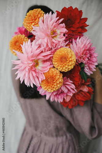 Beautiful woman holding autumn flowers bouquet in rustic room. Atmospheric aesthetic image. Florist in linen dress hiding behind beautiful colorful dahlias. Autumn season in countryside