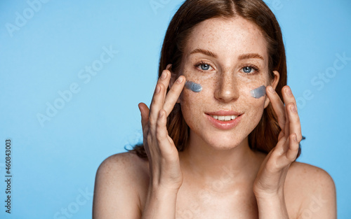 Portrait of redhead woman applying clay mask, skin care product on her face, smiling from pleasure, using cosmetic facial cream, standing over blue background