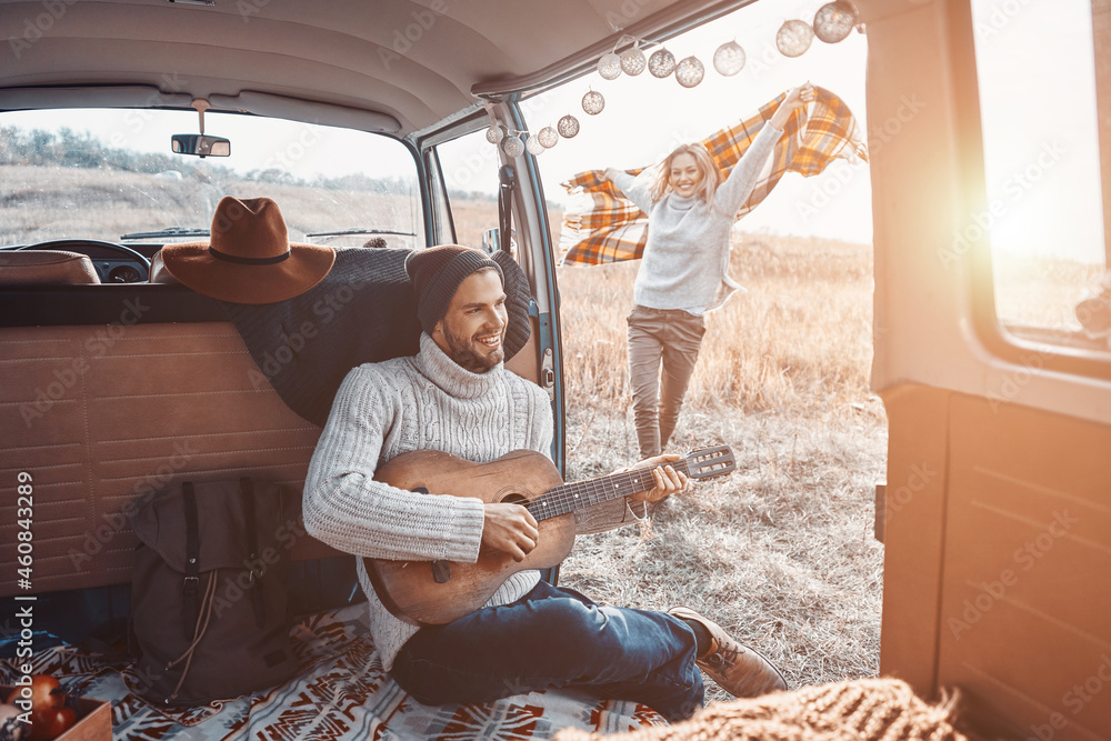 Handsome young man playing guitar for his girlfriend while spending time in motor home - obrazy, fototapety, plakaty 
