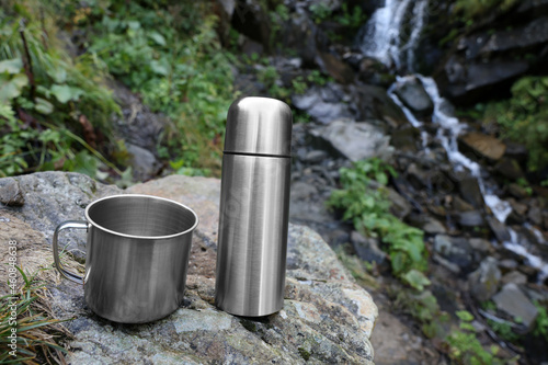 Cup and vacuum flask on rock near mountain waterfall. Space for text