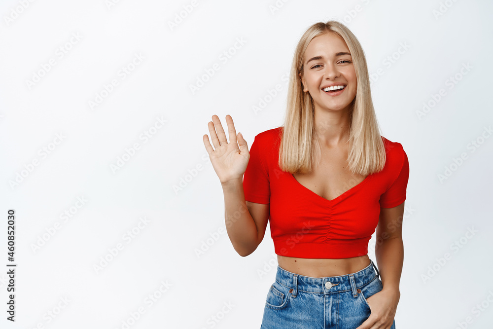 Friendly stylish woman saying hello, waving hand to greet you and smiling, standing in cropped top with jeans against white background Friendly stylish woman saying hello, waving hand to greet you and smiling, standing in cropped top with jeans against white background