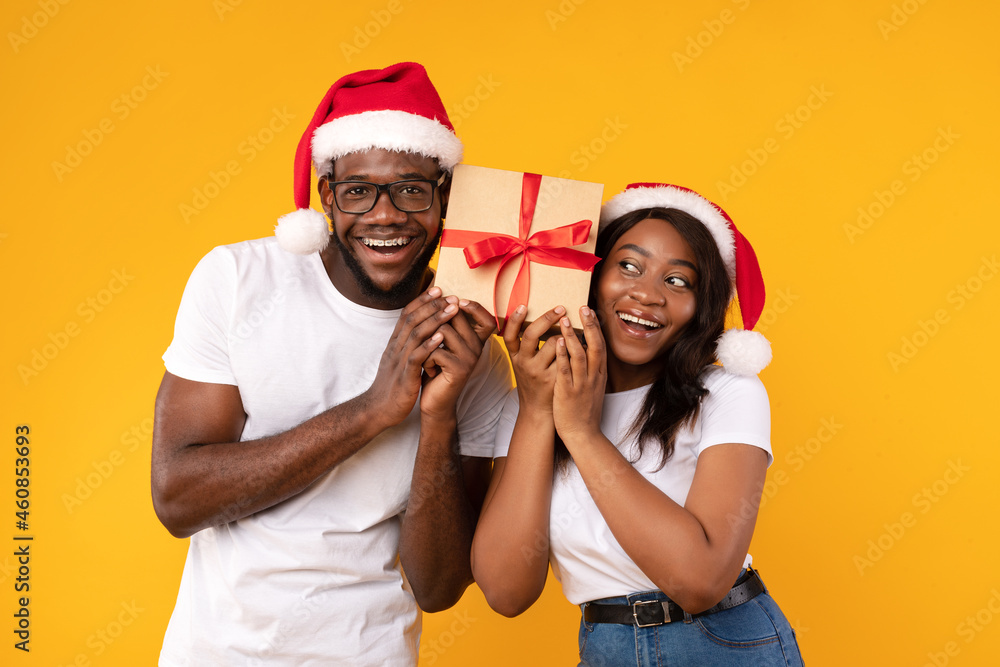 Black Couple Holding Christmas Gift Guessing What's Inside, Yellow Background Black Couple Holding Christmas Gift Guessing What's Inside, Yellow Background