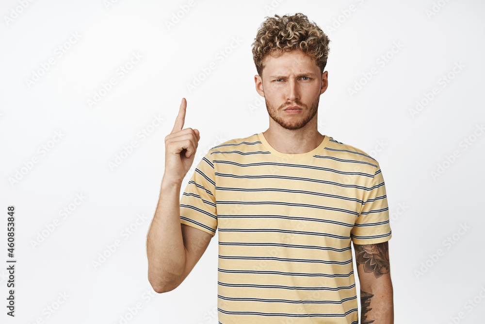Suspicious blond guy squinting, pointing finger up at something uncertain and doubtful, standing over white background in t-shirt Suspicious blond guy squinting, pointing finger up at something uncertain and doubtful, standing over white background in t-shirt